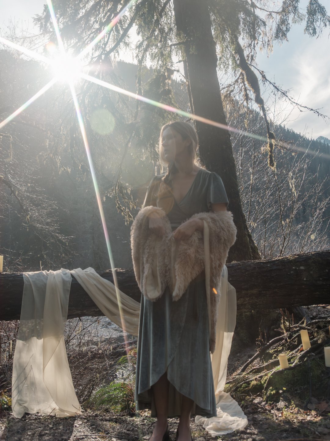 A woman standing outdoors in a forest near a large tree, with sunlight shining through the branches. She is wearing a gray dress and a fur coat draped over her arms, and there are white fabric and candles on the ground and a log nearby.