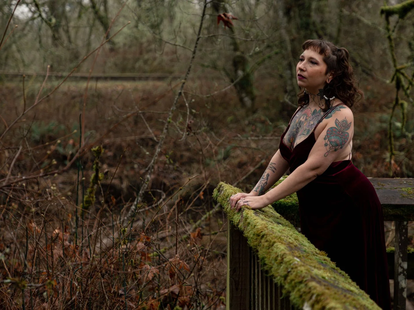 A woman with tattoos leaning on a moss-covered wooden railing outdoors in a forest.