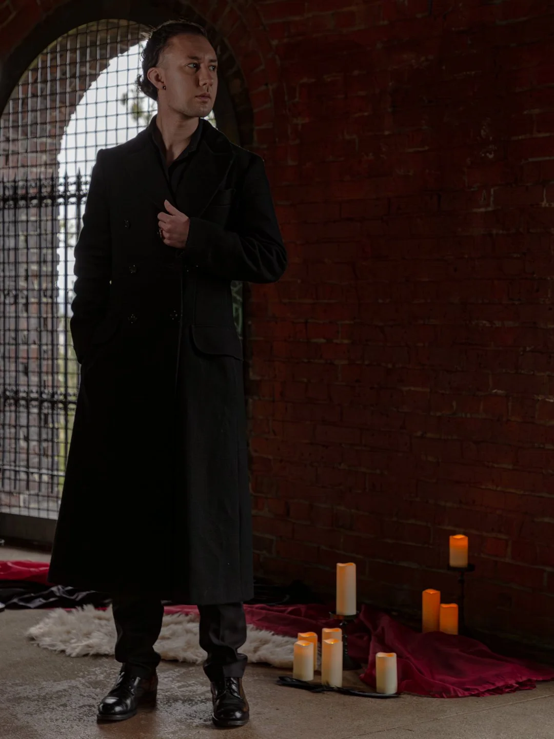 A man dressed in black stands in front of a brick wall with candles placed on the ground nearby, inside an arched structure with a metal gate in the background.