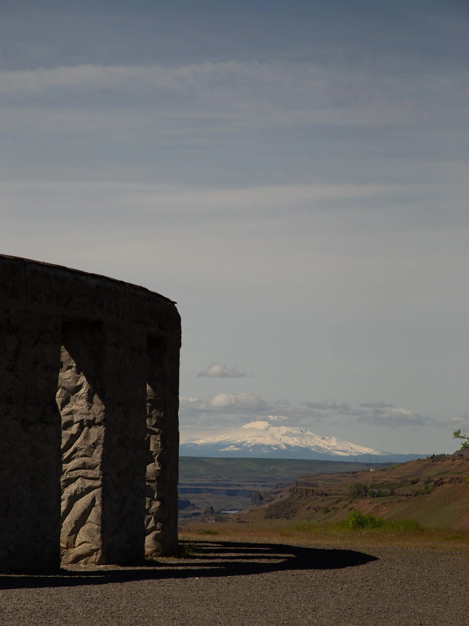 View of a bright blue sky with clouds, mountains with snow caps in the distance, and a landscape of green and brown rolling hills and valleys, seen through the gap of an old stone bridge or structure.