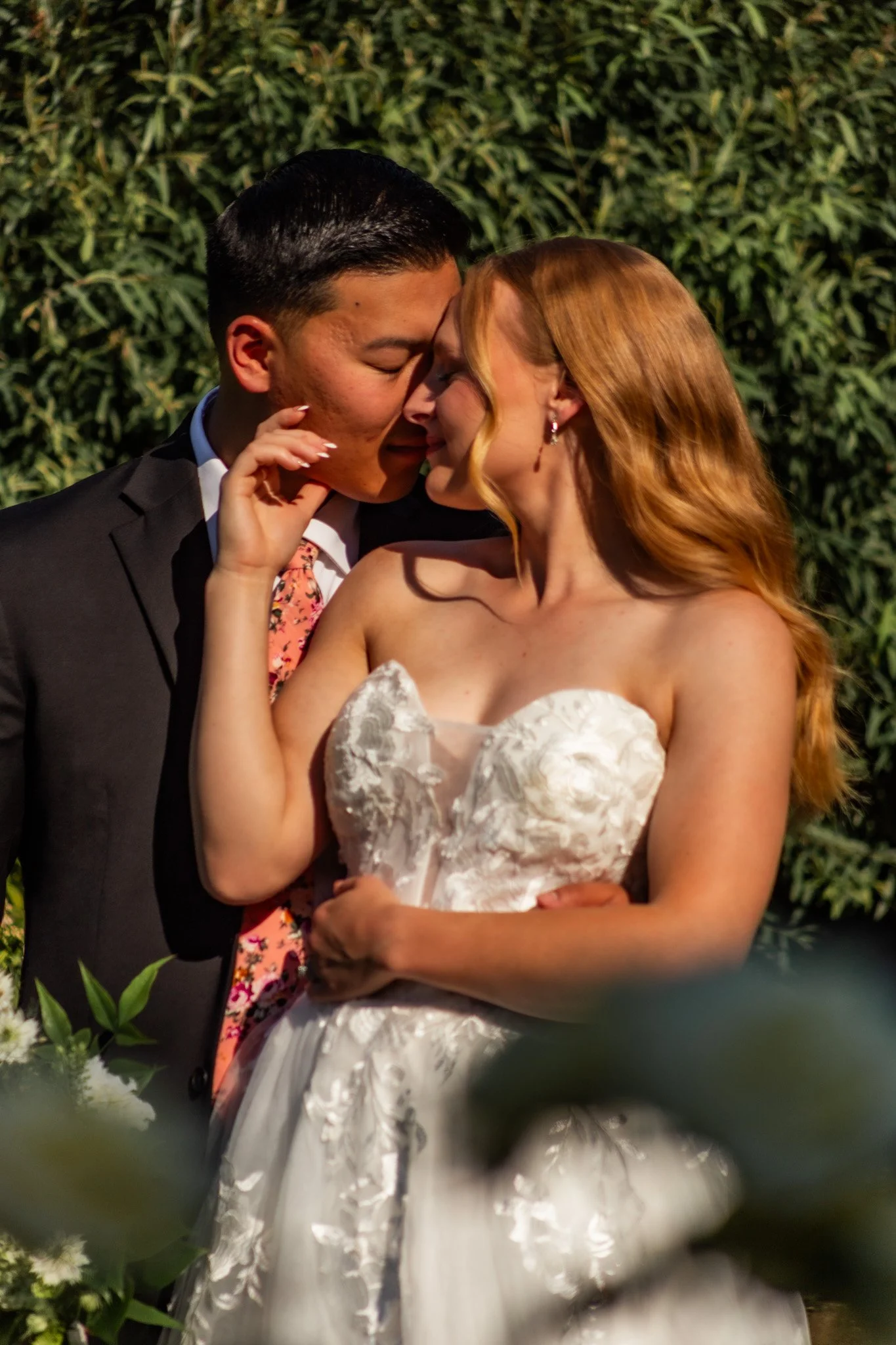 A bride and groom sharing a kiss at their wedding ceremony. The groom is wearing a black tuxedo and a floral tie, and the bride is in a white strapless wedding dress with floral embroidery. They are surrounded by greenery and floral arrangements.