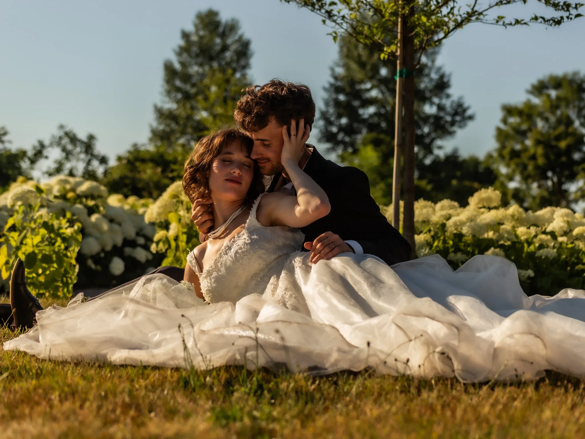 A couple is sitting on the grass in a field of white flowers, embracing each other and smiling. The man is wearing a dark suit, and the woman is wearing a white dress with a pearl necklace. Trees are visible in the background under a clear sky.