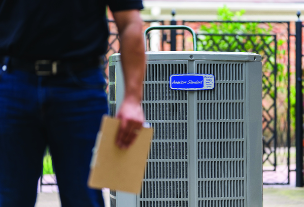 Close-up of a person holding a cardboard folder near a gray outdoor air conditioning unit labeled 'American Standard,' with a black metal fence and greenery in the background.