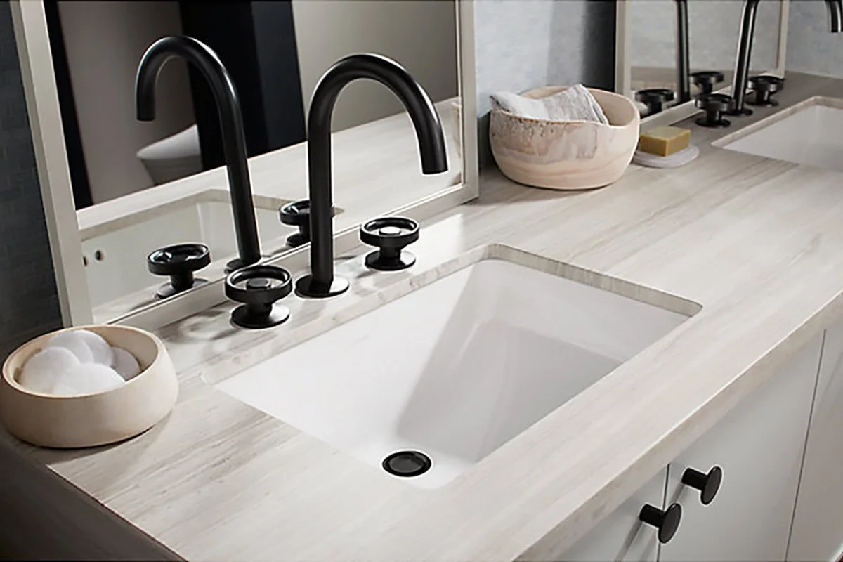 Modern bathroom sink with black faucet and handles, beige countertop, decorative bowls, soap, and a mirror.