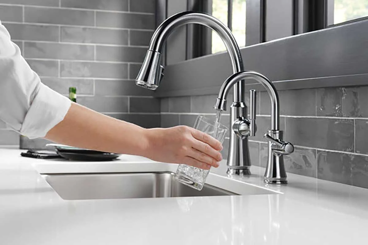 Person washing a glass under a kitchen sink faucet with a gray tiled backsplash.