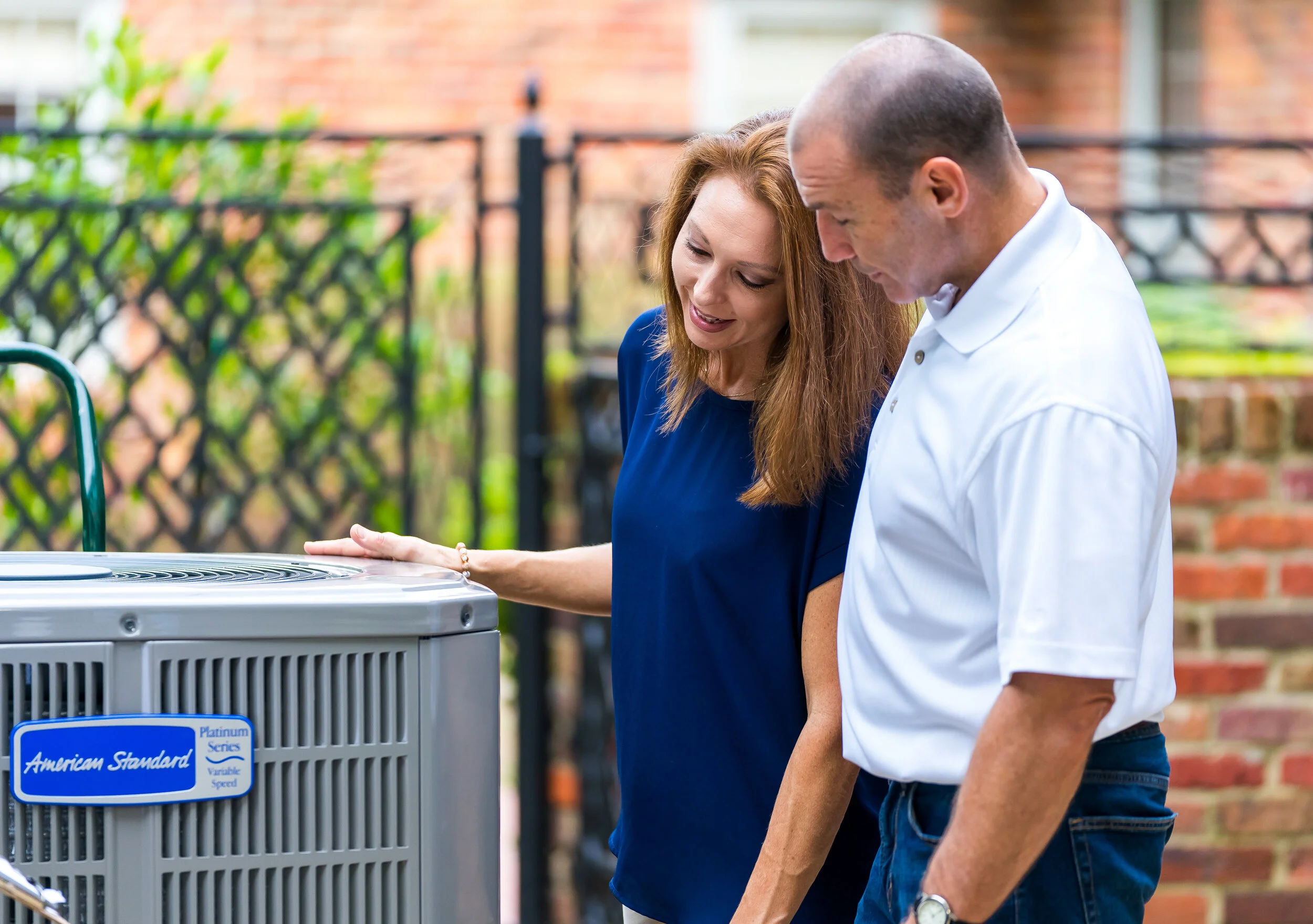 A man and a woman looking at an air conditioning unit outdoors on a patio with a brick wall in the background.