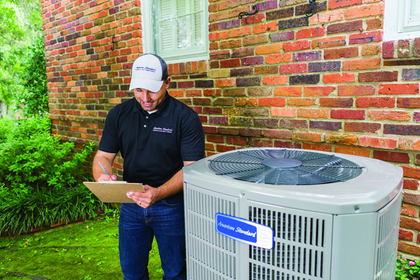 A man in a black polo shirt and white cap smiling and taking notes on a clipboard next to an outdoor air conditioning unit in front of a brick house.