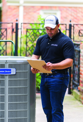 A man wearing a navy polo shirt and a cap, holding a clipboard, standing outdoors near an HVAC unit.