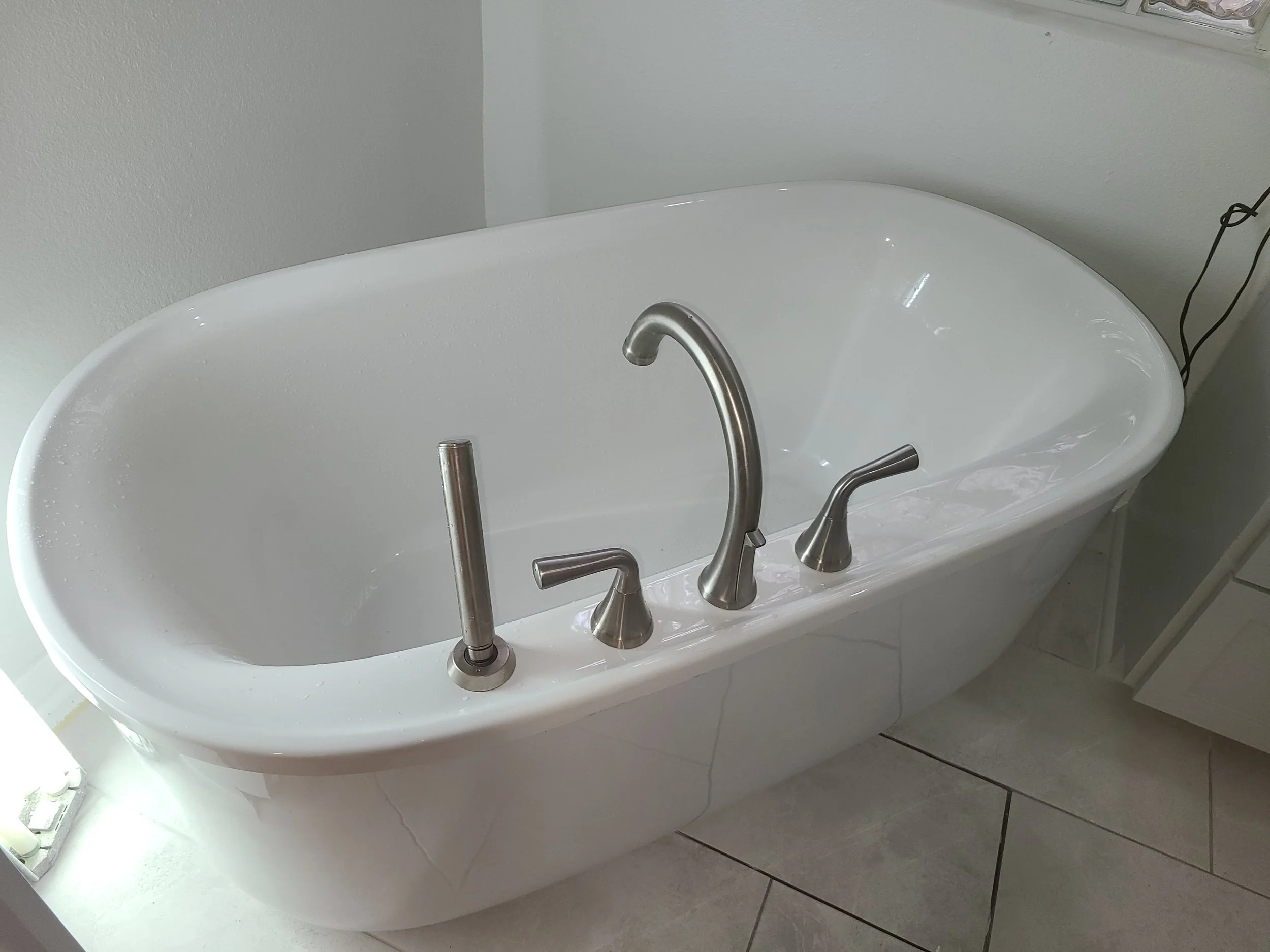 A white bathtub with chrome faucet and handles, set against a light gray wall in a bathroom. Two black electrical cords are visible on the right side.