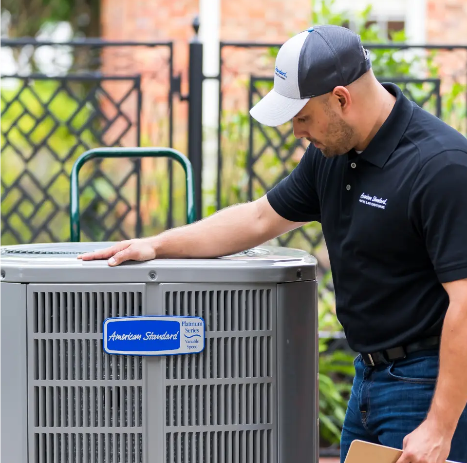 A man in a black polo shirt and baseball cap working on an American Standard air conditioning unit outdoors, with a black fence and green plants in the background.