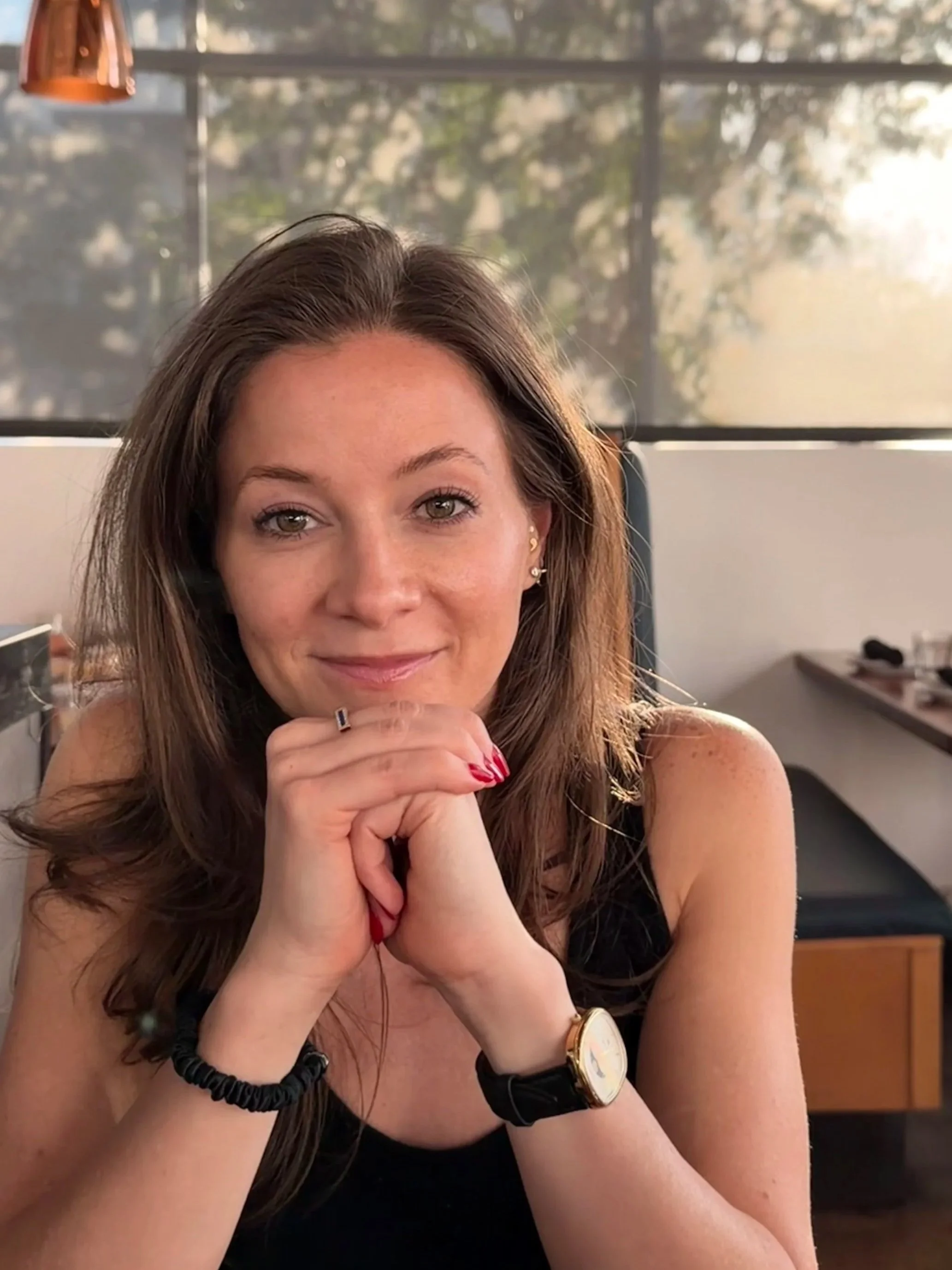 A woman with long brown hair smiling at camera, hands clasped near her chin, indoors with blurred background of windows and trees outside.
