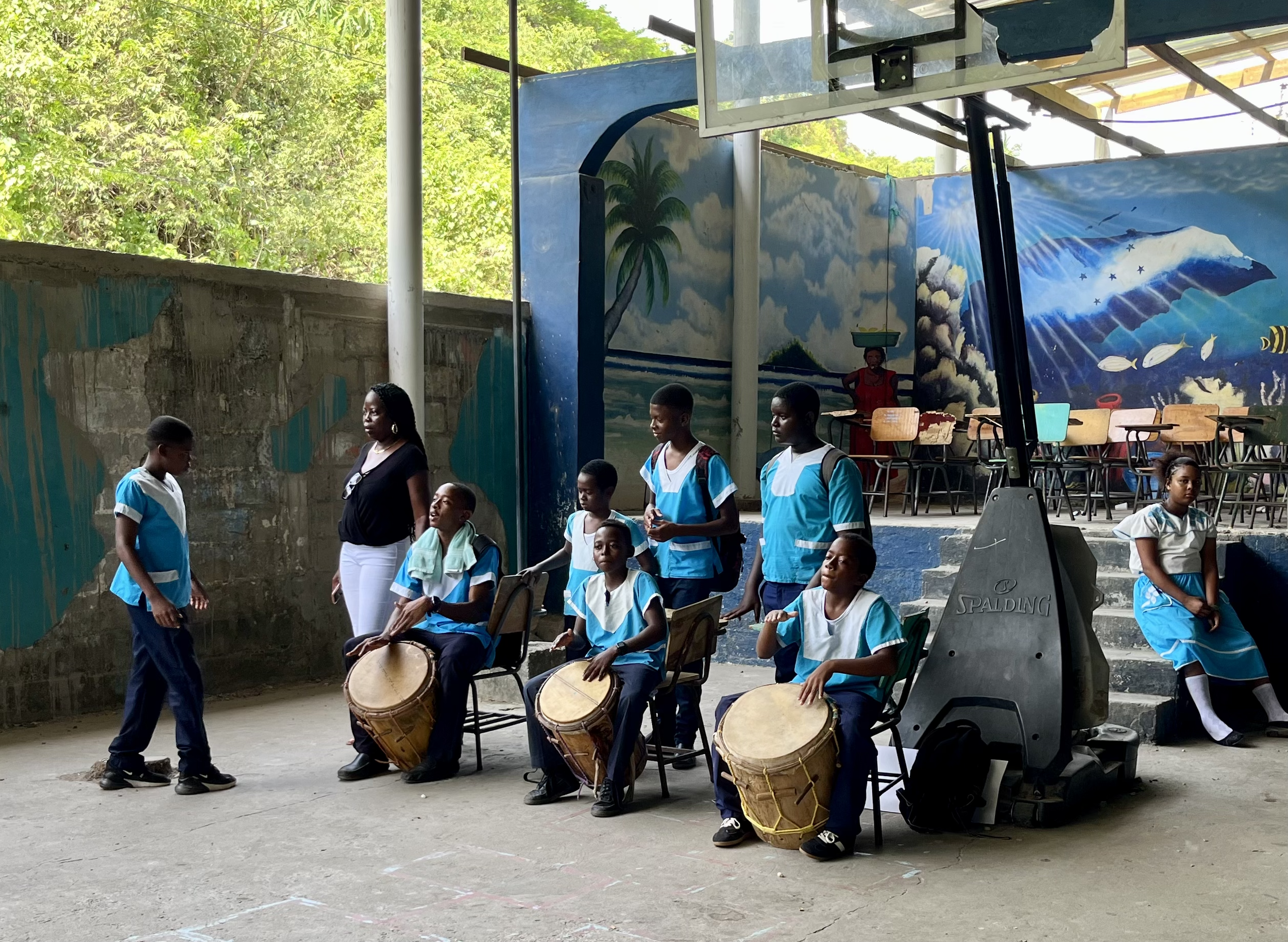 Garifuna students in teal school uniforms playing traditional drums in the covered outdoor court at the school in Punta Gorda, Honduras, with vibrant murals painted on the walls behind them