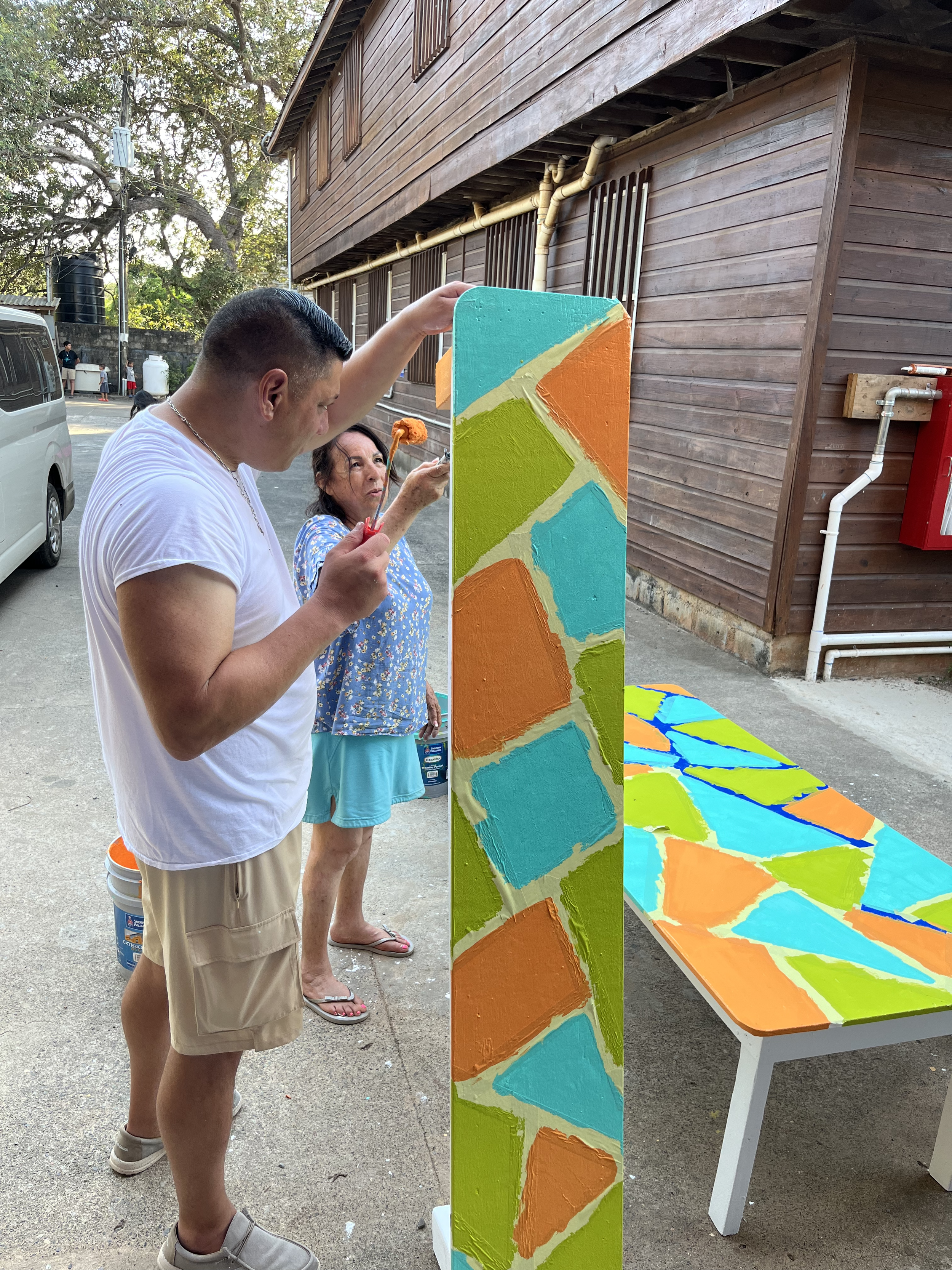 Two New City team members painting a colorful mosaic pattern in orange, teal, and green on a bench outside Sandy Bay Lighthouse Ministries in Roatán, Honduras