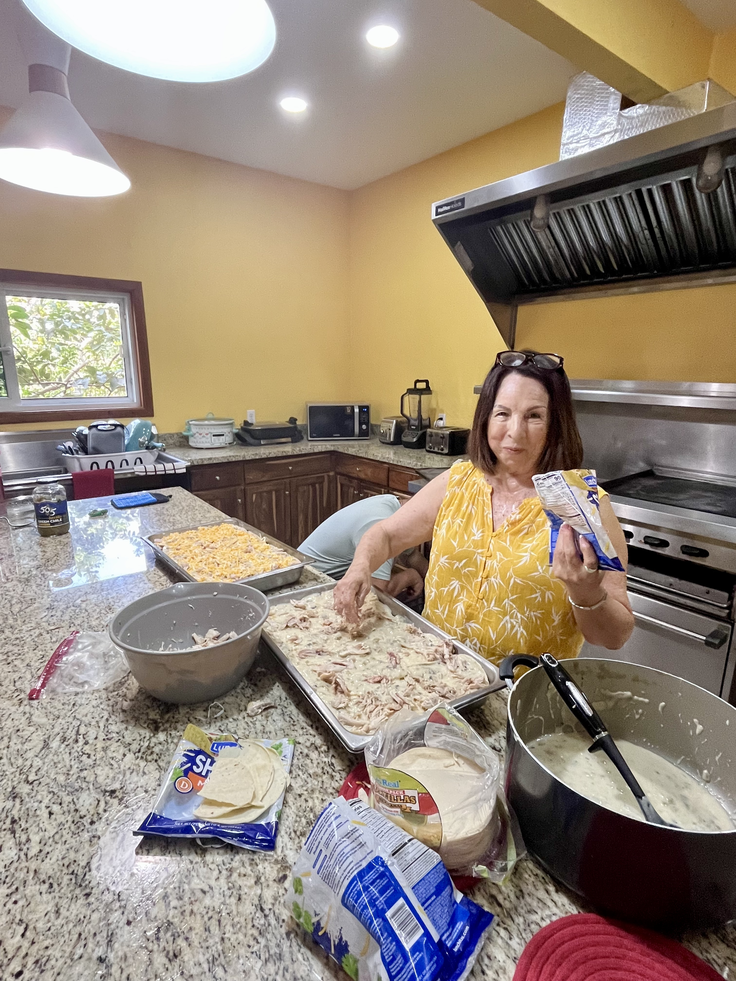 A New City team member assembling Green Chile Chicken Enchiladas in a commercial kitchen in Roatán, Honduras, with trays of shredded chicken and tortillas spread across the counter