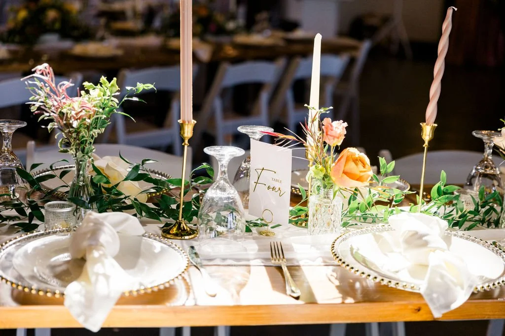 Elegant table setting with white plates, gold-rimmed chargers, silverware, napkins, pink and white candles, glass vases with pastel flowers, and greenery, for a formal event.