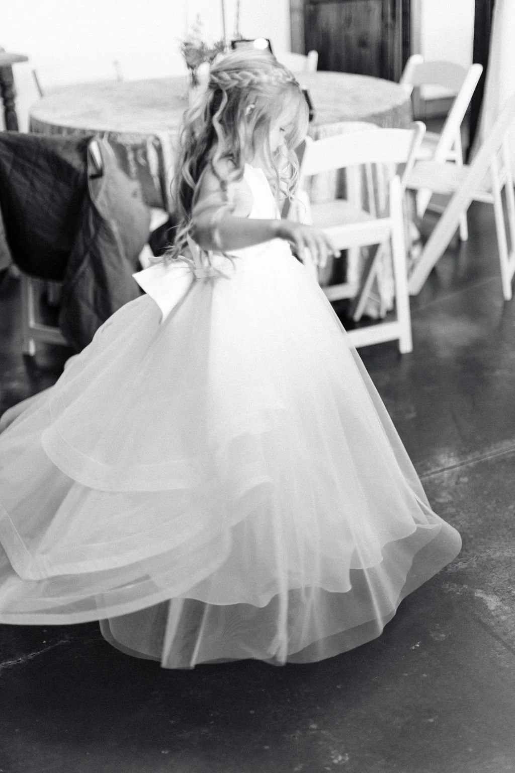 A young girl in a white dress twirling in a room with wooden chairs and a round table in the background.