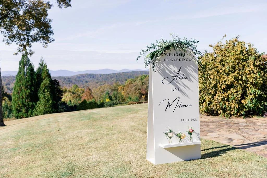 Wedding welcome sign with floral decoration on top, placed on grass with trees and mountains in the background.