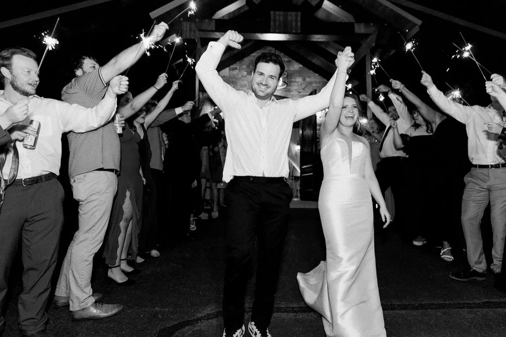 A newlywed couple walks through a celebration with guests holding sparklers, inside a modern mountain  venue.