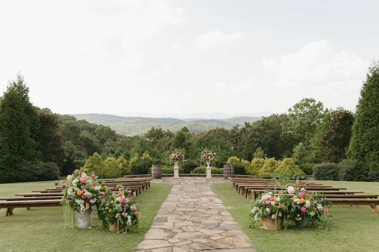 Outdoor wedding ceremony setup with flower arrangements, wooden benches, a stone aisle, and scenic mountain and forest backdrop.