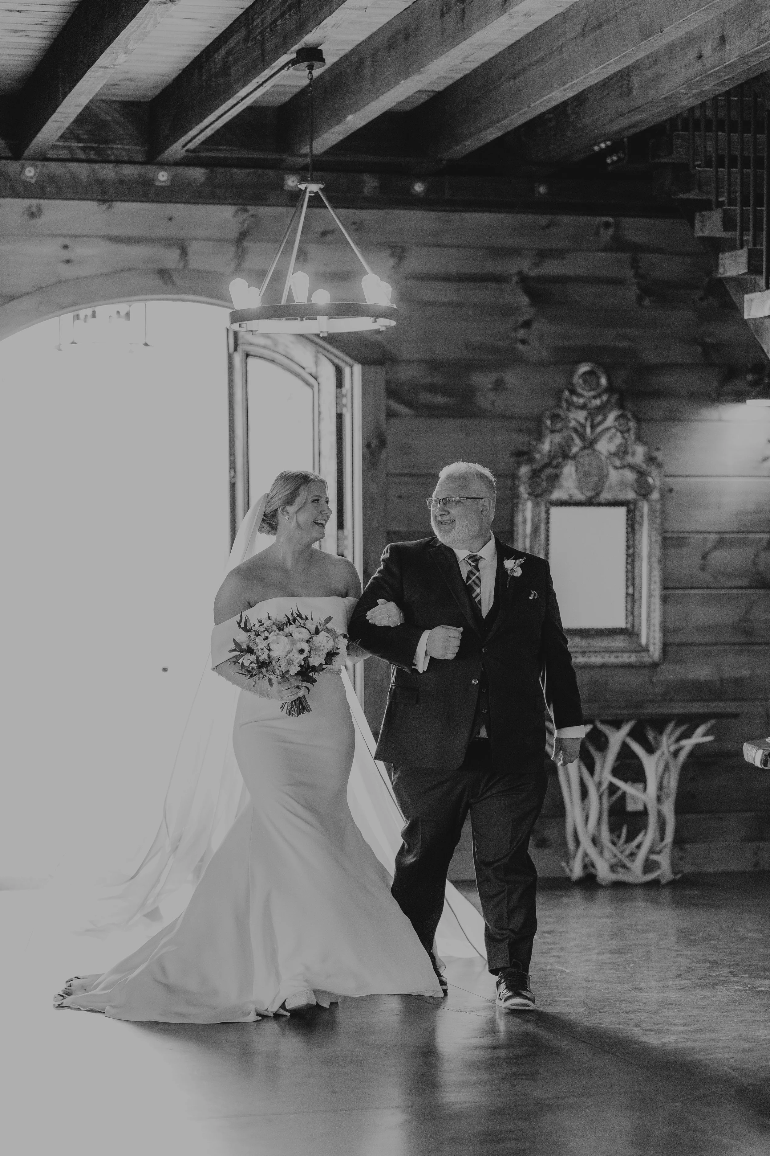 A bride in a wedding gown holding a bouquet of flowers is walking down the aisle with an older man, likely her father, in a suit, inside a wooden venue during her wedding.