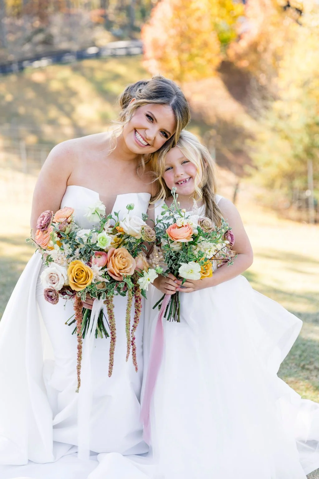 A bride and a young girl dressed in white, holding bouquets of colorful flowers, standing outdoors with trees and fall foliage in the background.