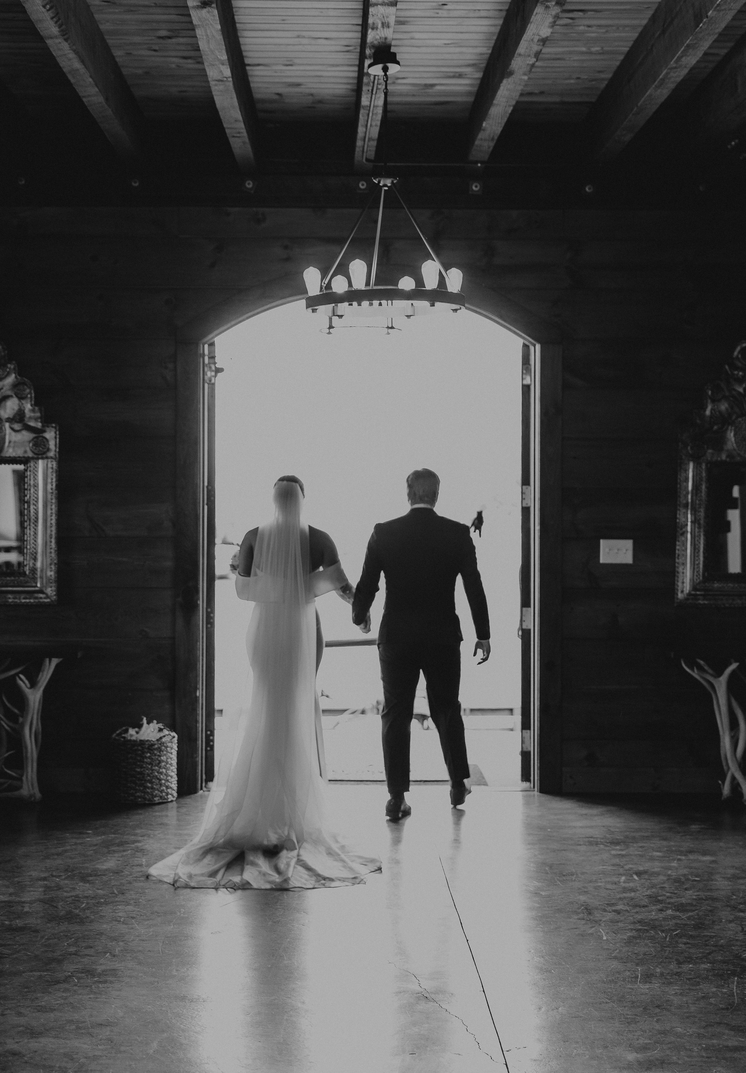A bride and groom holding hands, walking toward an open barn door with bright outdoor light.