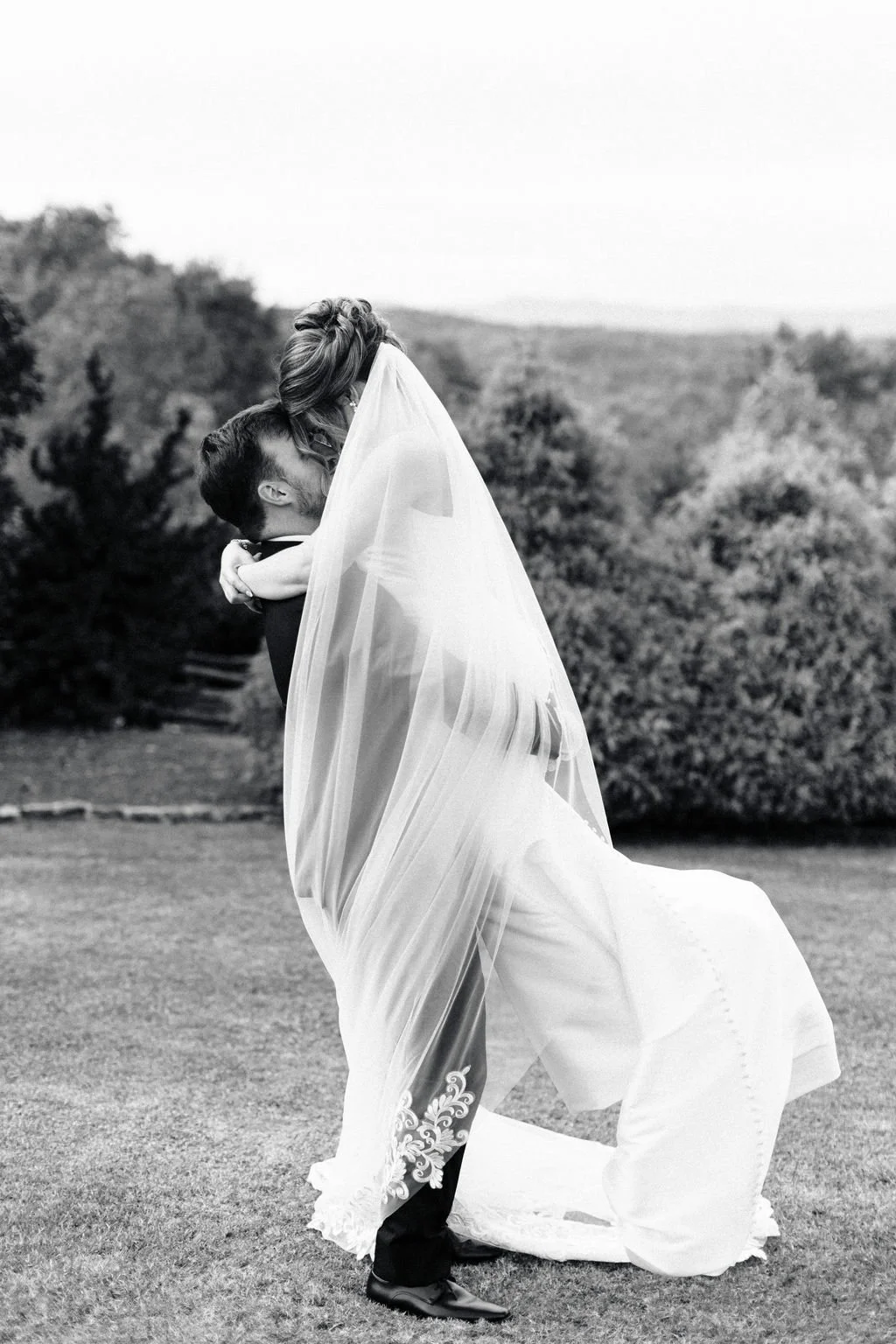 A black and white photo of a bride lifted by a groom outdoors, with trees and open land in the background.
