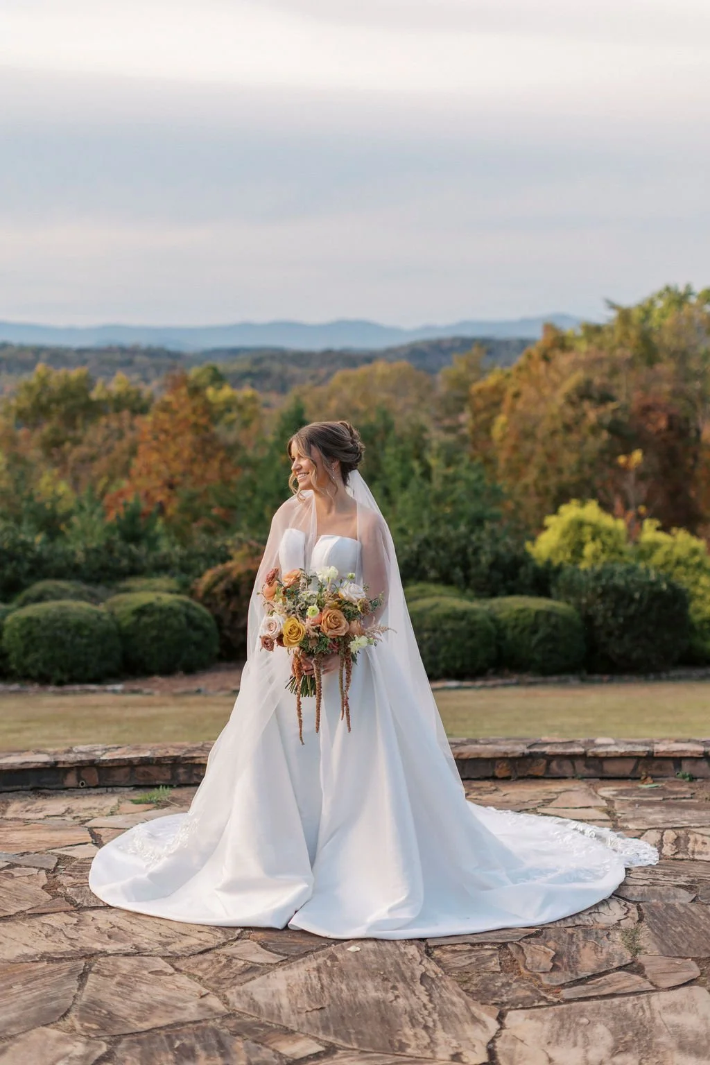 A bride in a white wedding gown holding a bouquet of roses and greenery, standing outdoors on a stone patio with trees and mountains in the background.