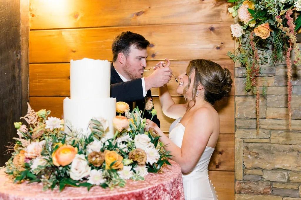 Bride and groom feeding each other cake during their wedding reception, with a decorated wedding cake and floral arrangements in the foreground.