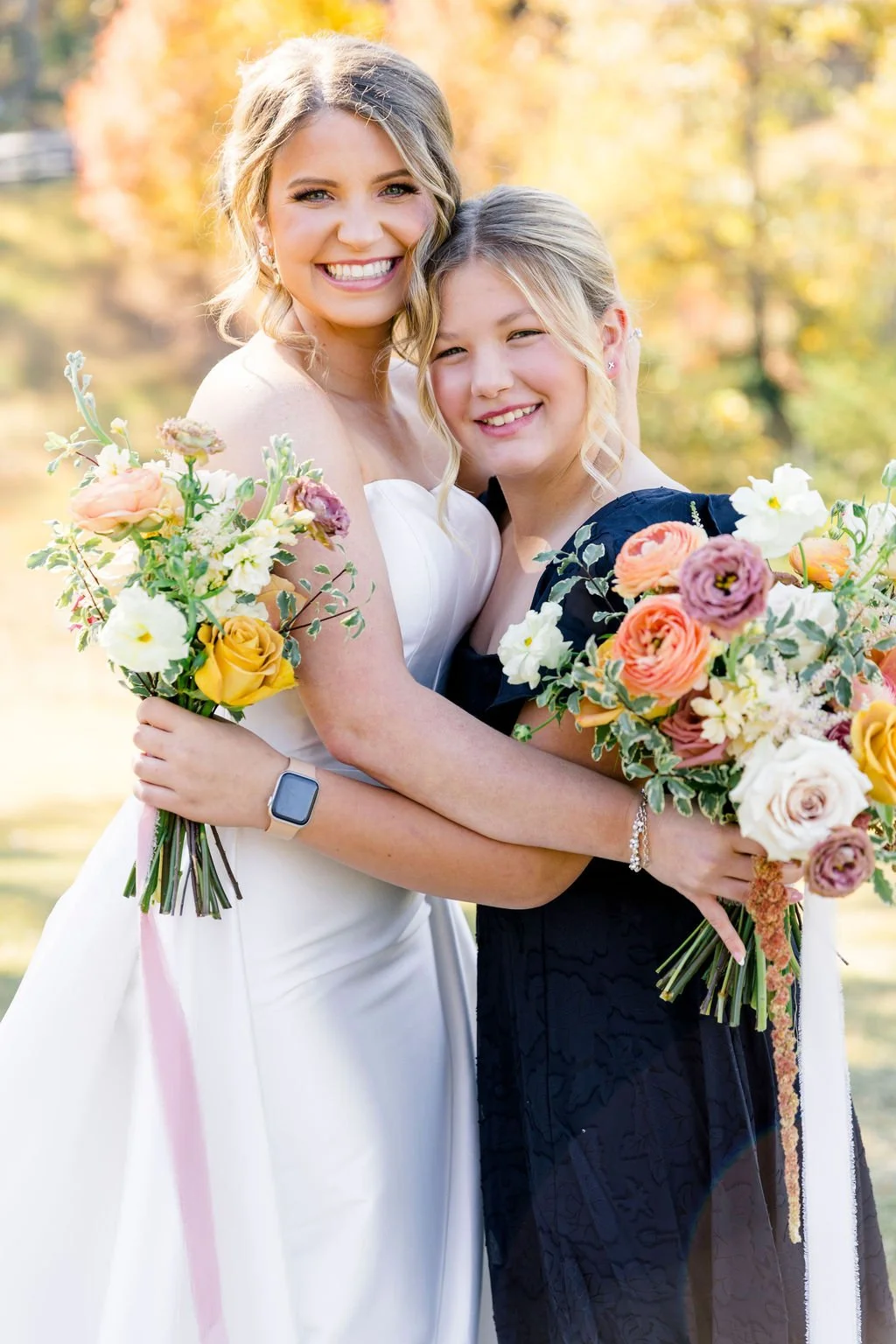 Two women, one in a white dress and the other in a black dress, smiling and hugging outdoors, holding colorful bouquets of flowers during autumn.