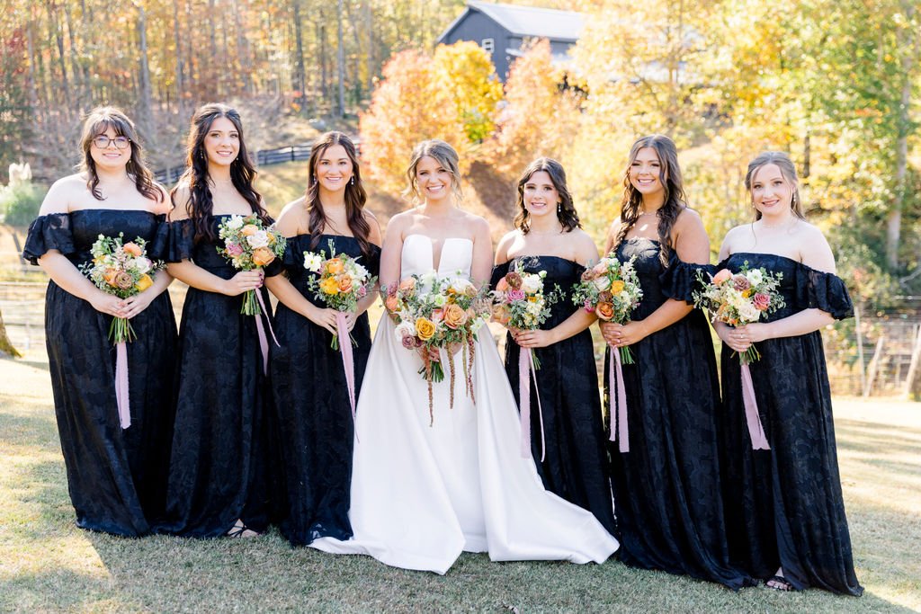 Bridal party consisting of seven women standing outdoors in fall, with trees and a building in the background. One woman wears a white wedding dress, the others wear matching black off-the-shoulder gowns. They hold bouquets of flowers with pink, peac