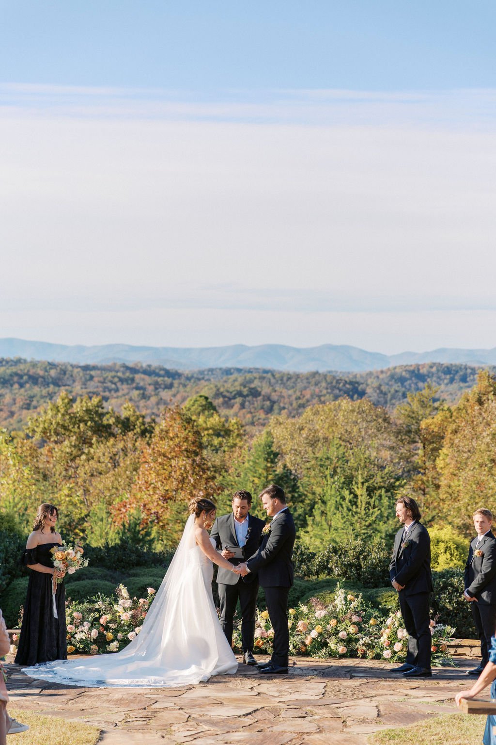 A wedding ceremony taking place outdoors in a scenic mountain setting with a couple exchanging vows, surrounded by bridesmaids and groomsmen, and floral arrangements.