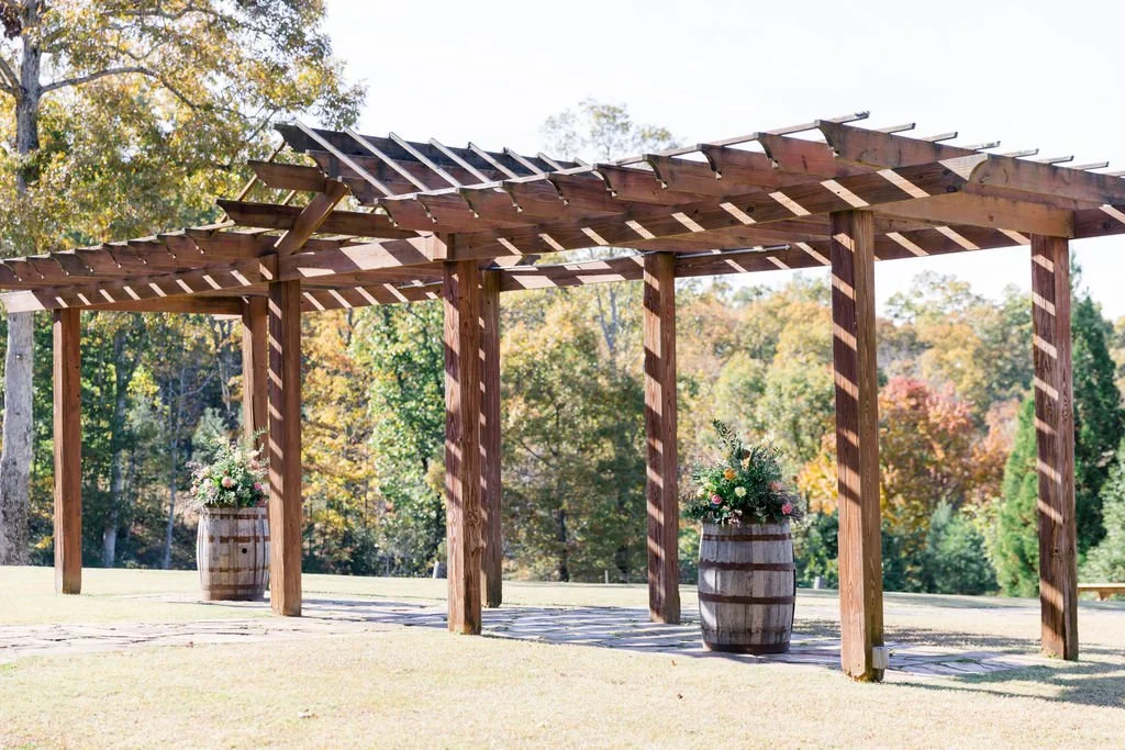 Wooden pergola with flower arrangements in barrels on a grassy field, surrounded by trees with fall foliage.