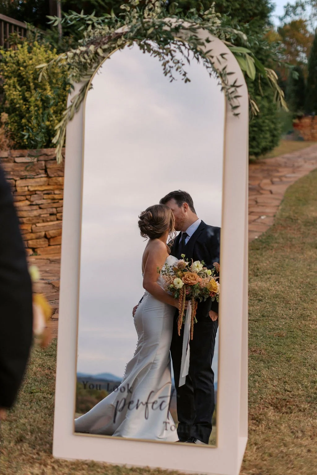 A bride and groom share a kiss during their wedding ceremony, seen through a tall, decorative mirror with greenery and flowers at the top.