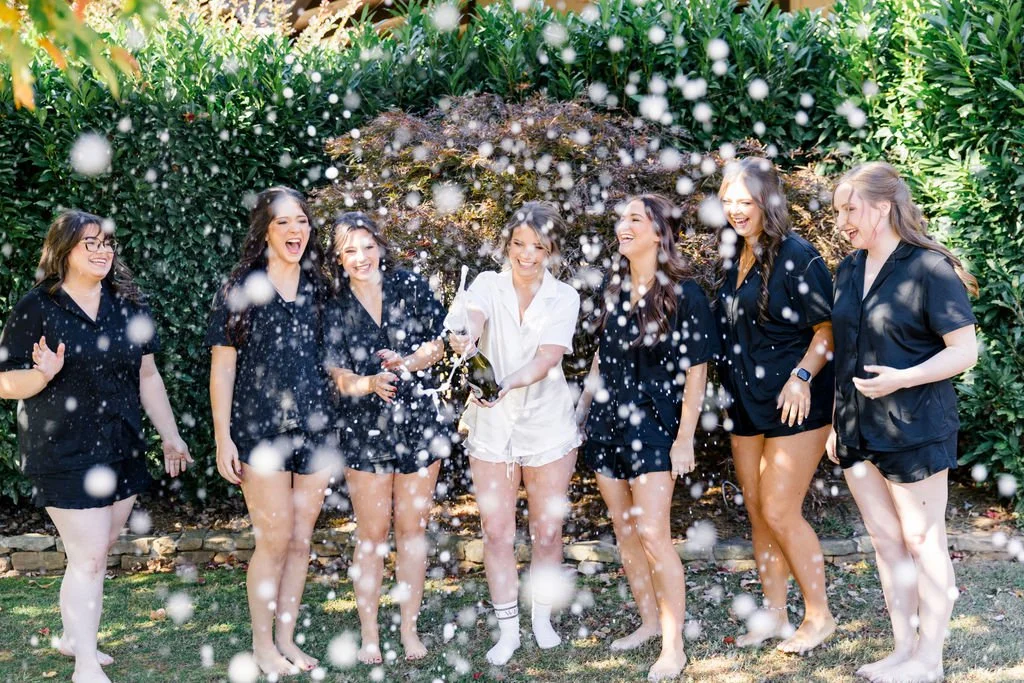 Group of women celebrating outdoors, popping champagne, surrounded by greenery, with bubbles floating around.