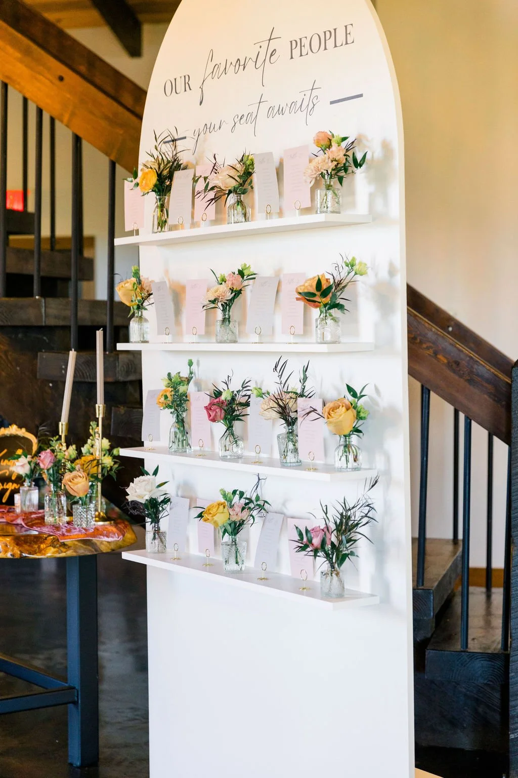 A white display board with the words 'Our favorite people, your seat awaits' written on it, decorated with small flower arrangements in glass jars with pink cards in each.