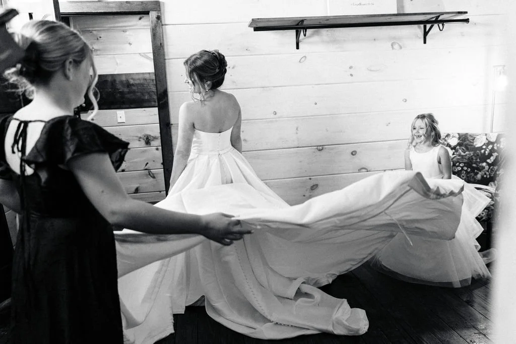 A woman in a wedding dress is being assisted by two girls, one holding the dress, in a refined farmhouse room with a mirror and a shelf, while another girl sits on a floral couch smiling.