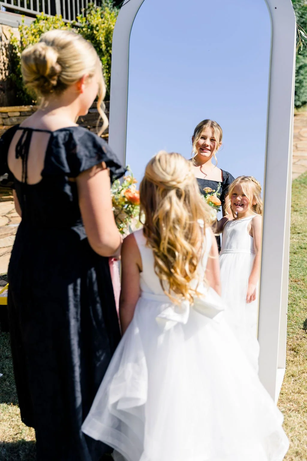 A young girl in a white dress with a large bow on the back stands outdoors in front of a large mirror, smiling at her reflection. She is surrounded by an older girl and a woman, likely at a wedding or formal event. The scene is bright and sunny with 