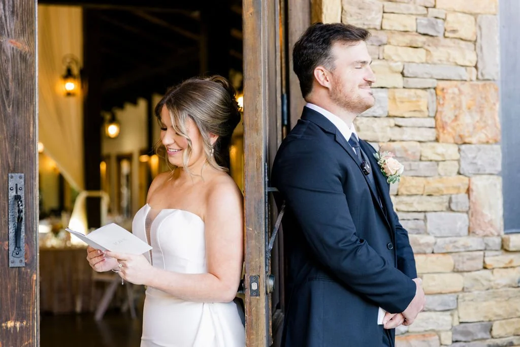 Bride in a white wedding gown reading a note, groom in a black suit leaning against a stone wall at a wedding venue.