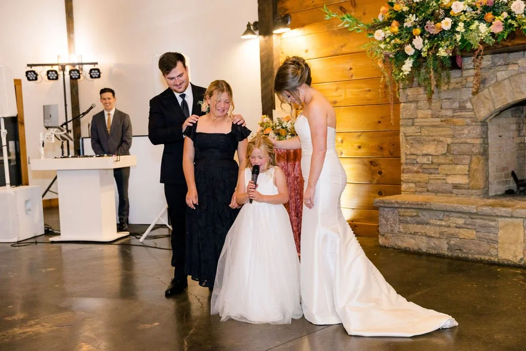 Bride in a white wedding gown standing with a young girl in a white dress holding a microphone, a man in a suit and tie, and a girl in a black dress, at a wedding reception with a rustic stone fireplace and floral arrangement.