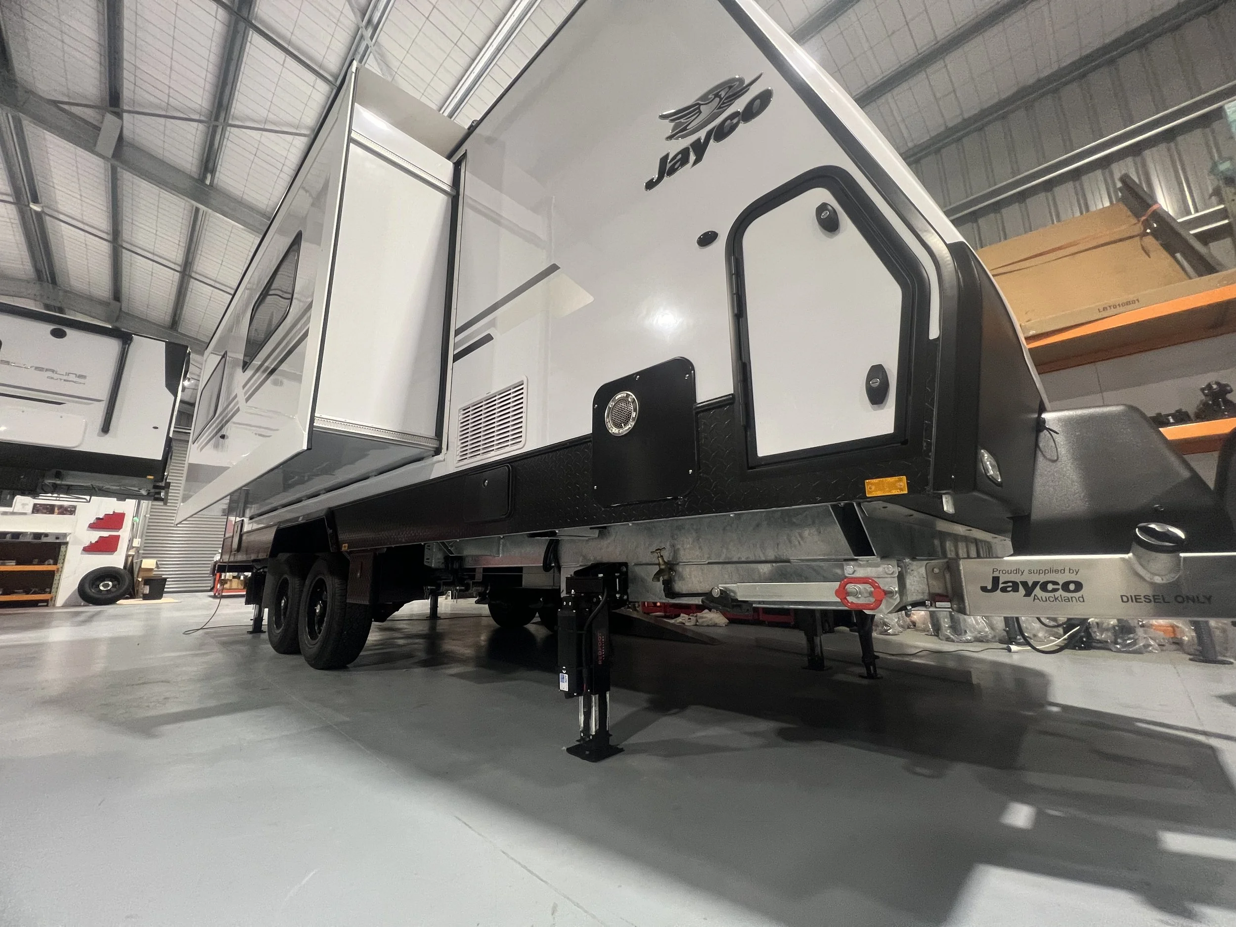 A Jayco travel trailer inside a warehouse, with black wheels and a white exterior, seen from a low angle.
