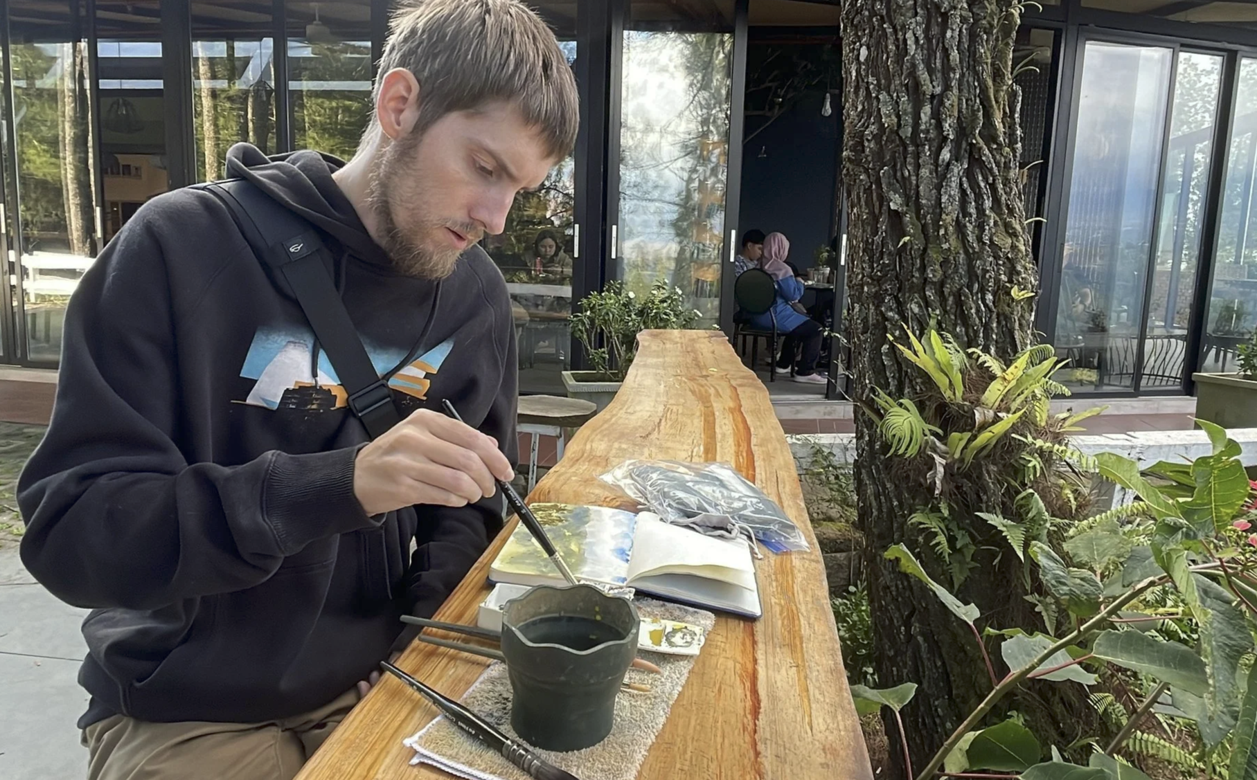 A man with a beard and gray hair, wearing a black hoodie, is sitting at a long wooden table outdoors, painting or drawing in an open sketchbook with a small brush. There are various painting supplies on the table, including a cup of dark-colored liquid and several brushes. Behind him, large glass windows reveal an indoor area with people sitting at tables, and there is a large tree with rough bark near the table, surrounded by green plants.