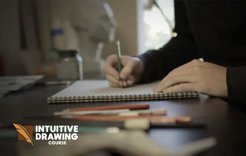 A person is drawing or writing in a spiral notebook on a table with colored pencils and pens in front of them. The setting appears to be a creative workspace, with a focus on artistic activity.