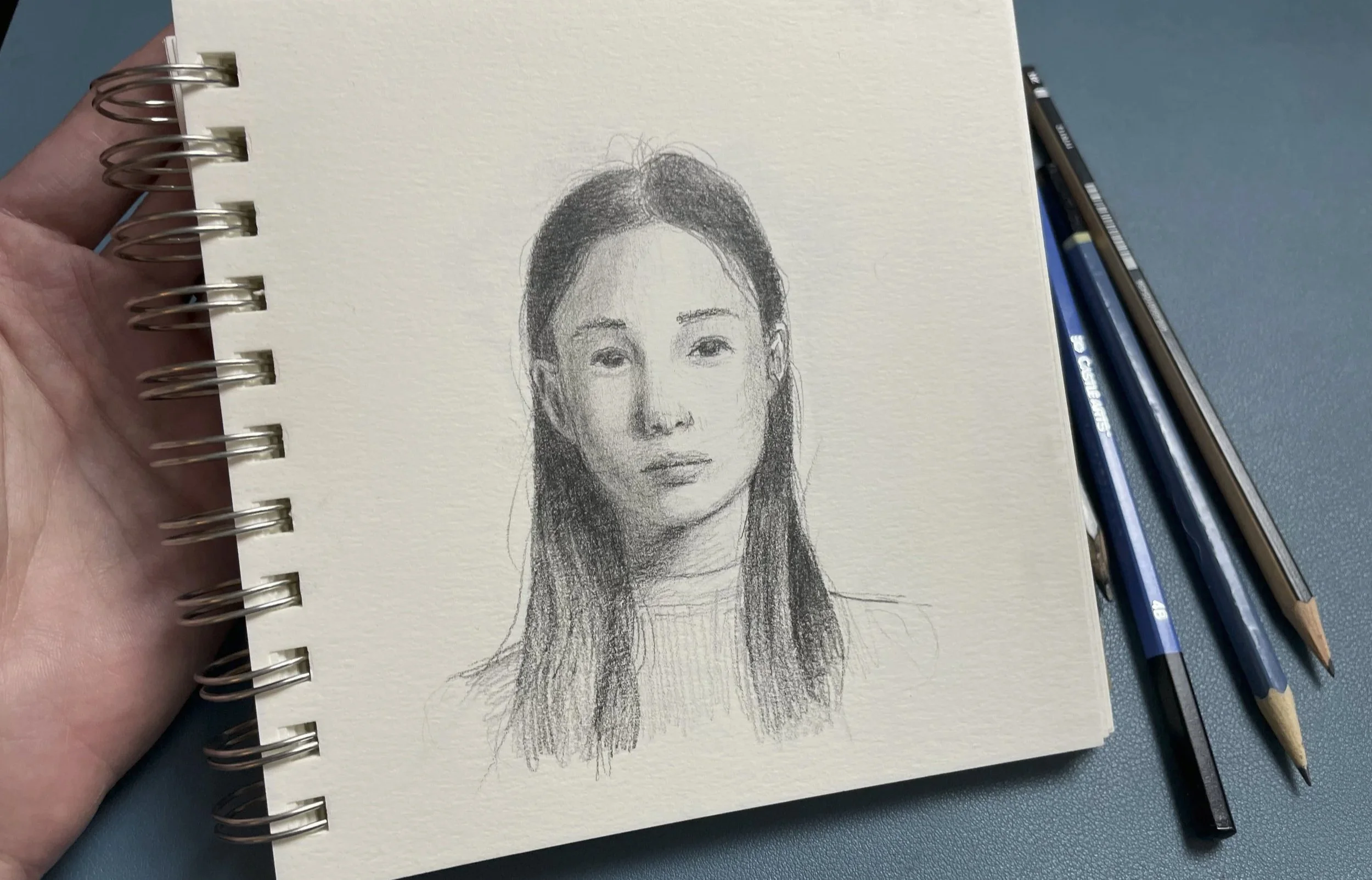 A hand holding a sketchbook with a detailed pencil portrait of a woman with long, dark hair on the page, with three pencils lying on a blue surface beside the sketchbook.