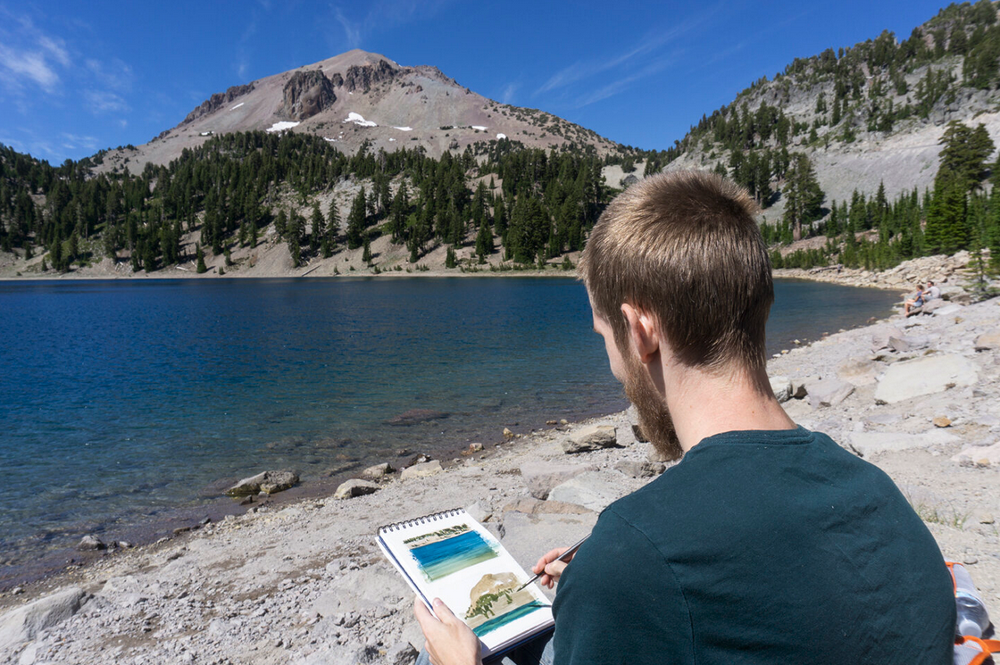 A man sitting by a mountain lake, painting on a sketchpad, with forested mountains and a clear blue sky in the background.