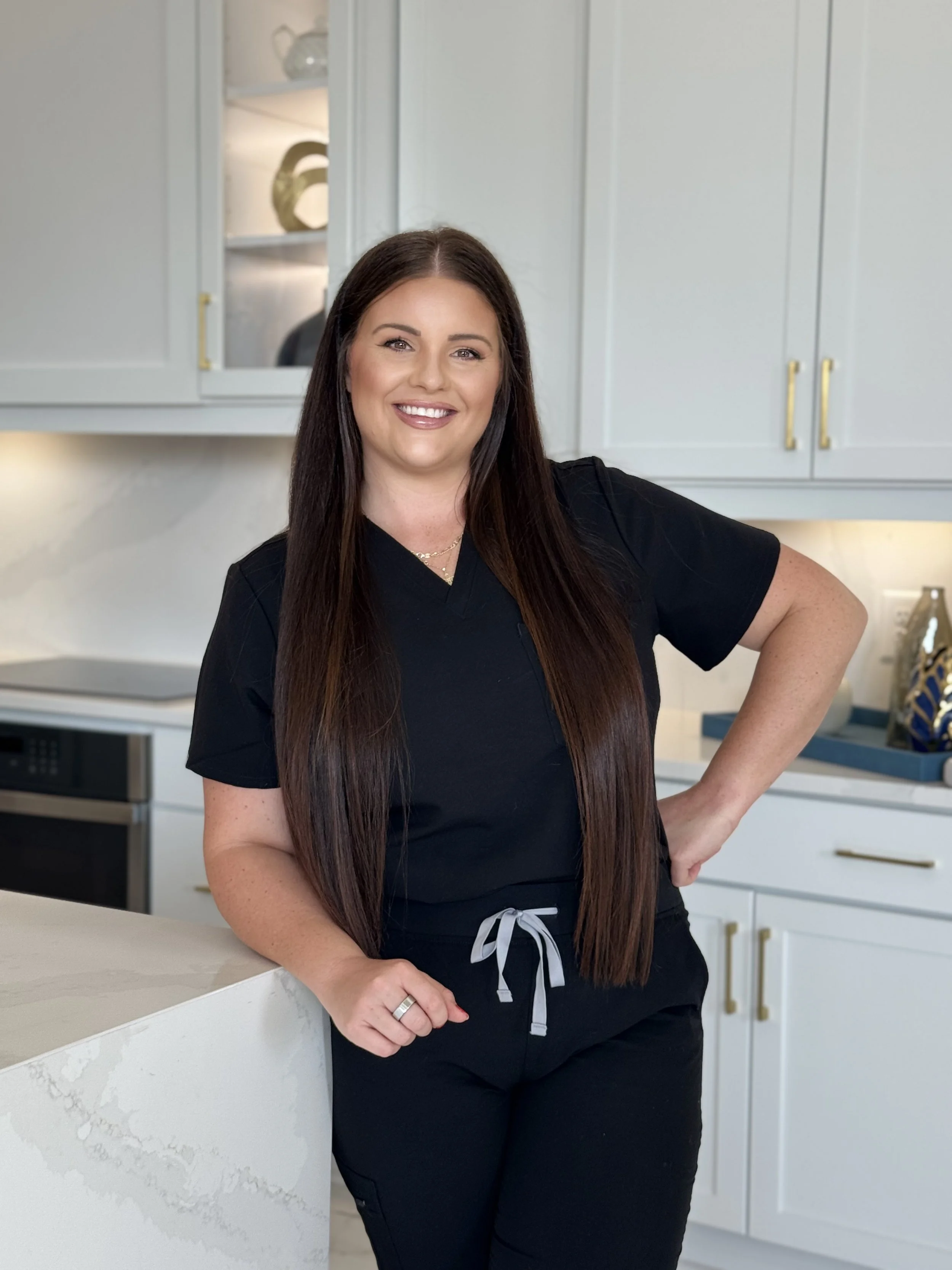 A woman with long dark hair smiling outdoors during sunset, wearing a white top with thin straps.