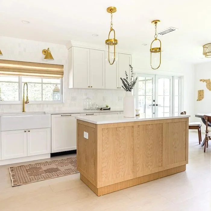 Modern kitchen with white cabinets, a wooden island, gold light fixtures, and a window with a beige shade.
