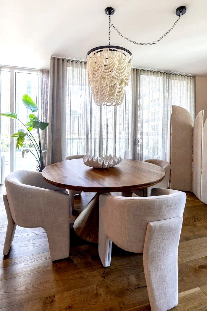 Round wooden dining table with four upholstered cream chairs, a decorative white ceramic bowl in the center, a large window with sheer curtains, and a chandelier made of hanging white beads.