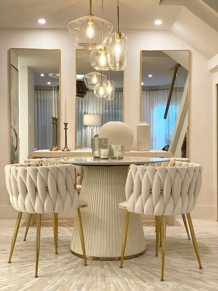 Elegant dining area with a round table, four plush white chairs with gold legs, decorative vases, candle holders, and a modern chandelier with glass globes, framed by three vertical mirrors on the wall.
