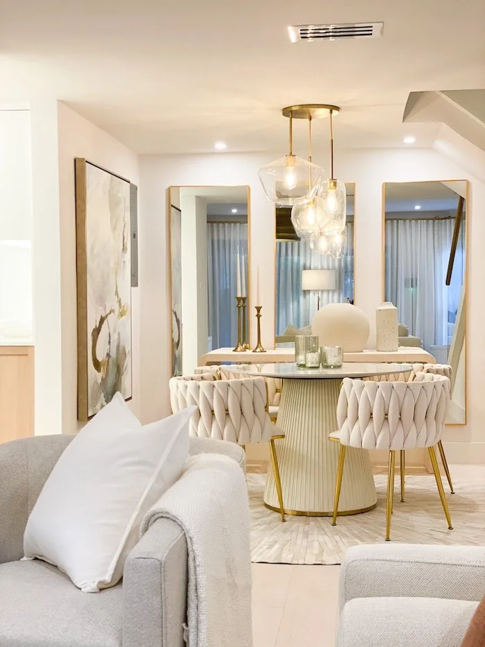Elegant dining area with a round marble table, upholstered chairs with gold legs, and decorative vases and candlesticks, reflected in large mirrors behind.